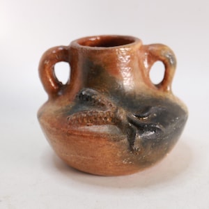 May include: A small, round, terracotta-colored clay vessel featuring two arched handles on the sides. The surface has a smooth, glazed finish with mottled patches of dark brown and black shading, and a raised, textured relief of a corn cob.