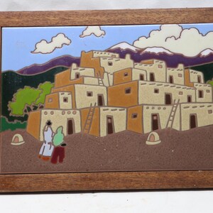 May include: A rectangular ceramic tile art piece framed in dark brown wood, depicting a multi-level adobe pueblo structure with ladders, two figures in the foreground, green trees, and snow-capped purple mountains under a blue sky with white clouds.