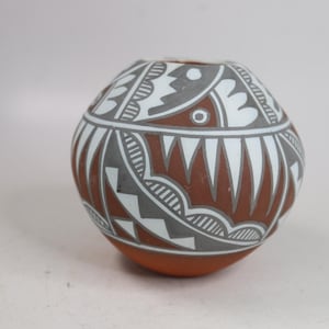 May include: A round terracotta pottery vessel featuring a matte finish with intricate hand-painted geometric patterns in white and dark gray. The design includes repeating triangular shapes, fine lines, and circular motifs encircling the body.