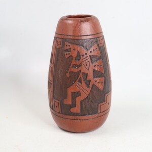 May include: A terracotta-colored vase with a tapered cylindrical shape, featuring dark brown etched geometric patterns and a central stylized figure playing a flute. The surface has a textured appearance with horizontal bands framing the design.