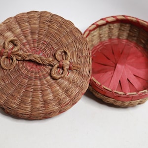 May include: A round woven basket with a matching lid. The basket features natural brown fibers and red accents. The lid has a central braided handle with decorative loops on each end. The interior base of the basket is painted a solid red color.