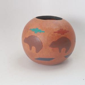 May include: A round, terracotta-colored vase with a dark opening. The vase is decorated with brown bear silhouettes, turquoise and red geometric shapes, and a blue stripe. The design has a Native American aesthetic.