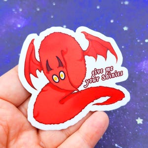 May include: A red sticker featuring a cartoonish devil-like creature with yellow eyes and small horns. The creature has bat-like wings and the text "give me your shinies" in a handwritten font.