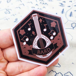 May include: A sticker with a brown and black design featuring a lute, musical notes, and the word "BARD" on a banner. The sticker has a white border and a hexagonal shape.