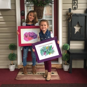 May include: Two young girls stand in front of a door holding signs that say "My First Day of 1st Grade!" and "My First Day of Pre-K!" The signs are framed in pink and purple and have watercolor backgrounds.