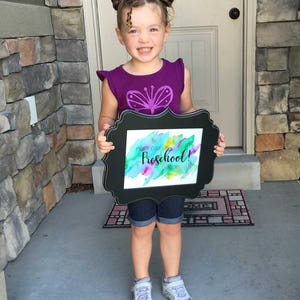 May include: A young girl with pigtails holds a framed sign that says "First Day of Preschool! 2018" in black lettering on a watercolor background. The sign is in a black frame with a decorative border. The girl is wearing a purple shirt with a butterfly design and denim shorts.