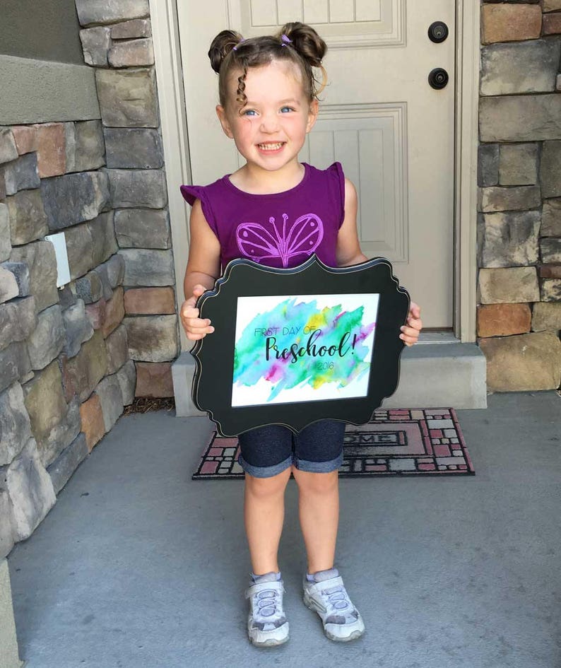 May include: A young girl with pigtails holds a framed sign that says "First Day of Preschool! 2018" in black lettering on a watercolor background. The sign is in a black frame with a decorative border. The girl is wearing a purple shirt with a butterfly design and denim shorts.