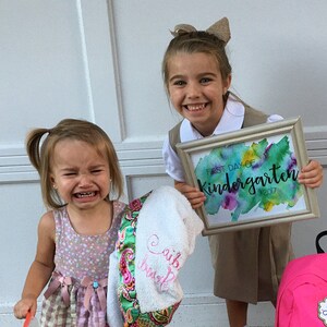 May include: Two young girls, one smiling and holding a framed sign that says "First Day of Kindergarten 2017" and the other crying, standing in front of a white door. The girl holding the sign is wearing a khaki skirt and a white shirt with a brown bow in her hair. The girl crying is wearing a pink and white dress and a white towel with pink embroidery.