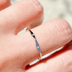 May include: A delicate gold ring with a subtle, textured design and a small, sparkling gemstone. The ring is worn on a finger, showcasing its minimalist style and elegant simplicity. The ring's slender band and the gemstone's sparkle create a refined aesthetic.