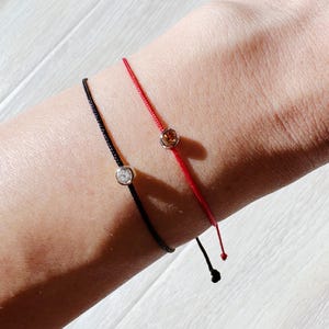 May include: Two delicate string bracelets, one black and one red, each with a small, sparkling gemstone. The black bracelet has a clear stone, while the red bracelet has a warm-toned stone. The bracelets are simple and elegant.