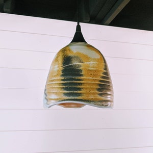 May include: A ceramic pendant light fixture with a black cord and a bell-shaped shade. The shade is glazed in shades of brown, yellow, and blue, with a textured, striated pattern.