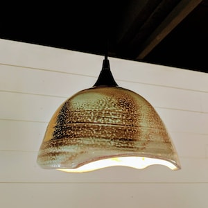 May include: A ceramic pendant light fixture with a brown and green glaze. The light is on and the light shines through the uneven edges of the shade.