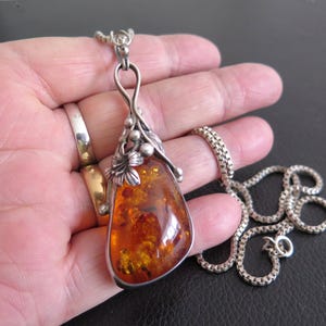 May include: A silver pendant necklace featuring a large, teardrop-shaped amber stone. The amber is a warm, translucent orange with internal patterns. The pendant has a decorative silver bail with leaf and berry details, suspended from a silver chain.