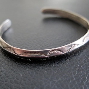 May include: A silver cuff bracelet with an etched design. The open bracelet features a repeating pattern of curved lines and small, triangular shapes. The metal has a slightly tarnished appearance, giving it a vintage look.