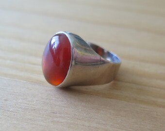 May include: A silver ring with a large, oval, reddish-orange gemstone. The ring has a thick band and a polished finish, showcasing the vibrant color of the stone. The gemstone is smooth and rounded, set in a simple, classic design.