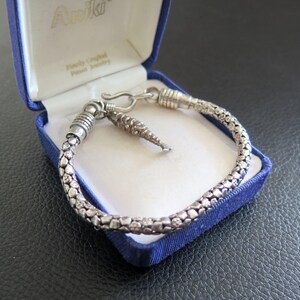 May include: A silver-toned bracelet with a textured, pebble-like surface and a small, dangling fish-shaped charm. The bracelet features a hook-and-eye clasp mechanism and is presented inside a blue, hinged jewelry box with the brand name Ariki.