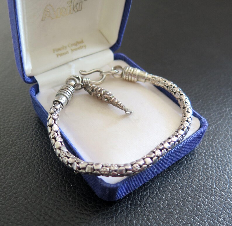 May include: A silver-toned bracelet with a textured, pebble-like surface and a small, dangling fish-shaped charm. The bracelet features a hook-and-eye clasp mechanism and is presented inside a blue, hinged jewelry box with the brand name Ariki.