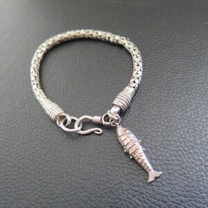 May include: A silver-toned bracelet featuring a woven, chain-link band with a textured, cylindrical clasp. A small, articulated fish-shaped charm hangs from a jump ring near the hook-and-eye closure, which secures the circular bracelet together.
