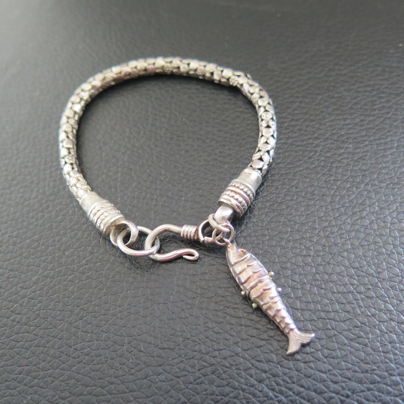 May include: A silver-toned bracelet featuring a woven, chain-link band with a textured, cylindrical clasp. A small, articulated fish-shaped charm hangs from a jump ring near the hook-and-eye closure, which secures the circular bracelet together.
