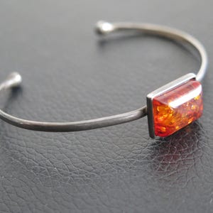 May include: Silver cuff bracelet with a rectangular amber stone. The amber has a warm, reddish-orange colour with internal patterns. The bracelet has rounded ends.