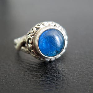May include: A silver ring with a large, round, vibrant blue gemstone. The ring has an ornate, detailed design around the gemstone and on the band. The gemstone is the focal point, set in a decorative bezel.