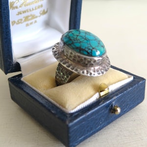May include: A silver ring with a turquoise stone set in a hammered silver band. The ring is in a blue velvet ring box.