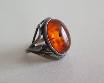 May include: A silver ring featuring a large, oval amber stone. The amber is a warm, translucent orange with dark inclusions. The ring has a detailed, leaf-like design on the band and around the stone, with a small ball accent.