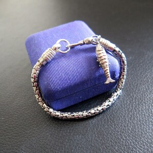 May include: A silver-toned bracelet featuring a textured, chain-link design with a metallic finish. The bracelet includes a small, dangling fish-shaped charm attached to a circular clasp mechanism, showcasing intricate detailing on the fish's body.