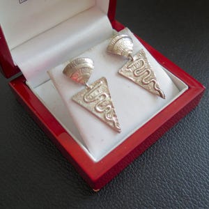 May include: A pair of silver-toned earrings displayed in a red jewelry box. Each earring features a textured, triangular pendant with a stylized, curved design. The earrings are presented on a white, cushioned interior.