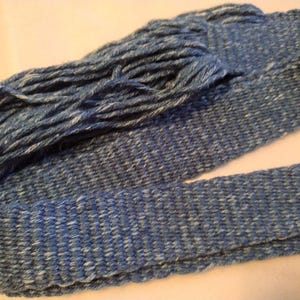 Puede incluir: Una longitud de hilo azul y blanco tejido, con una apariencia texturizada. El hilo está tejido firmemente en una banda plana, con un fleco de hilo suelto en un extremo. El efecto general es un aspecto rústico y hecho a mano.