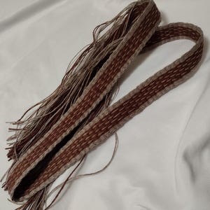 Puede incluir: Una correa tejida marrón y beige con flecos en un extremo. La correa está hecha de un material grueso y texturizado y tiene un patrón geométrico.