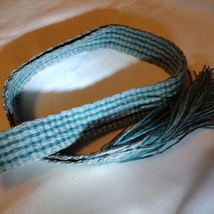 Puede incluir: Un cinturón tejido azul y gris con flecos en un extremo.