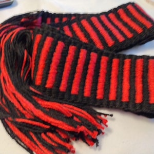 Puede incluir: Bufanda tejida en negro y rojo con un patrón de rayas. La bufanda tiene un fleco de hilo rojo en un extremo. Las rayas son verticales y espaciadas uniformemente. La bufanda está hecha de un material suave y texturizado.
