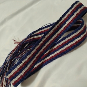 Puede incluir: Una tela tejida a rayas azul, blanca y roja con flecos en un extremo. La tela está doblada por la mitad y los flecos cuelgan.