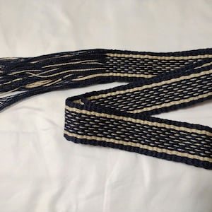 Puede incluir: Un cinturón tejido con flecos en un extremo. El cinturón es negro y crema con un patrón geométrico.