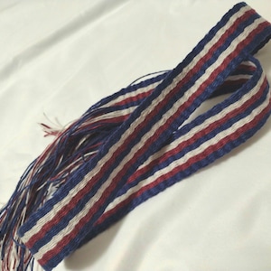 Puede incluir: Una tela tejida a rayas azules, blancas y rojas con flecos en un extremo.