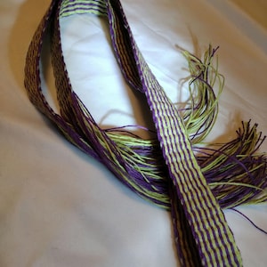 Puede incluir: Una correa tejida de color verde y morado con los extremos deshilachados. La correa está hecha de hilos finos y tiene un patrón a rayas.