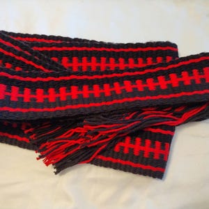 Puede incluir: Un cinturón tejido negro y rojo con flecos. El cinturón tiene un patrón geométrico de rayas rojas y negras.