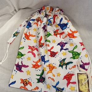 May include: A white drawstring bag with a colorful fish print. The bag has a white drawstring and a small tag that says "Beautifully and lovingly made".