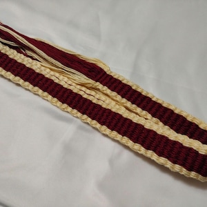 May include: A woven red and cream belt with fringe tassels. The belt is made of a thick, sturdy material and has a simple, elegant design.