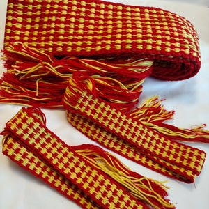 Puede incluir: Un cinturón tejido rojo y amarillo con flecos. El cinturón está hecho de un patrón de cuadros con cuadrados rojos y amarillos.