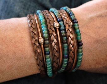 May include: A stack of bracelets with a braided leather band and turquoise blue glass beads. The bracelets have copper wire accents and small copper beads.