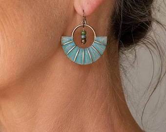 May include: A woman wearing turquoise and gold-colored fan-shaped earrings. The earrings have a half-circle design with a gold-colored hoop and small green beads. The woman is wearing a white blouse.
