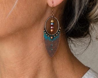 May include: A dangle earring featuring a twisted metal hoop at the top, suspended from a hook wire. The hoop contains three small, colourful beads. Below the hoop is a pointed, shield-shaped metal pendant with a teal and copper-toned patina finish.