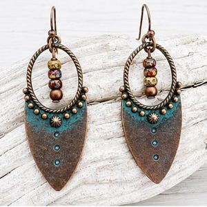 May include: A pair of dangle earrings featuring a copper-toned metal shield shape with a teal patina finish. Each earring includes a twisted circular frame, a central star-shaped stud, three small dots, and a vertical row of three multicolored beads.