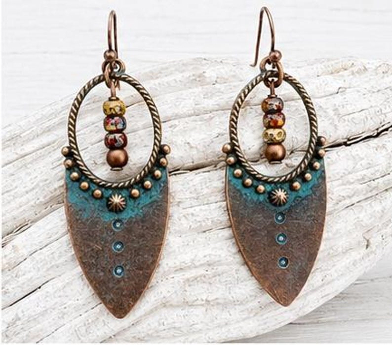 May include: A pair of dangle earrings featuring a copper-toned metal shield shape with a teal patina finish. Each earring includes a twisted circular frame, a central star-shaped stud, three small dots, and a vertical row of three multicolored beads.
