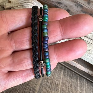 Puede incluir: Tres pulseras apiladas en una mano. La pulsera superior est&aacute; hecha de cuentas iridiscentes verdes y azules. La pulsera del medio es un cord&oacute;n de cuero negro con una cuenta plateada. La pulsera inferior es un cord&oacute;n de cuero negro con una cuenta plateada.