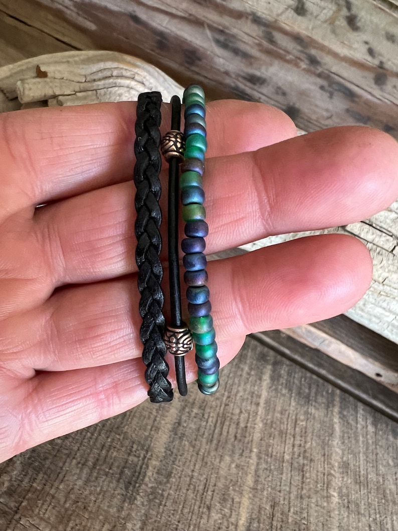 Puede incluir: Tres pulseras apiladas en una mano. La pulsera superior est&aacute; hecha de cuentas iridiscentes verdes y azules. La pulsera del medio es un cord&oacute;n de cuero negro con una cuenta plateada. La pulsera inferior es un cord&oacute;n de cuero negro con una cuenta plateada.