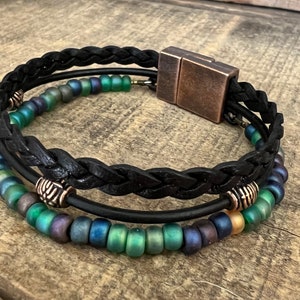 A black leather bracelet with a braided design and a magnetic clasp. The bracelet is adorned with iridescent green and blue beads.