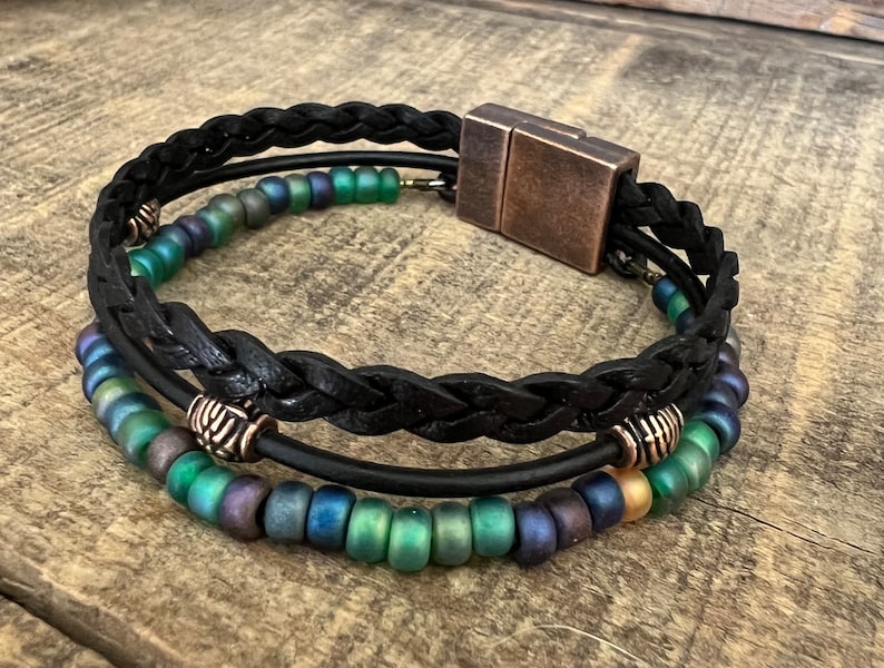 A black leather bracelet with a braided design and a magnetic clasp. The bracelet is adorned with iridescent green and blue beads.